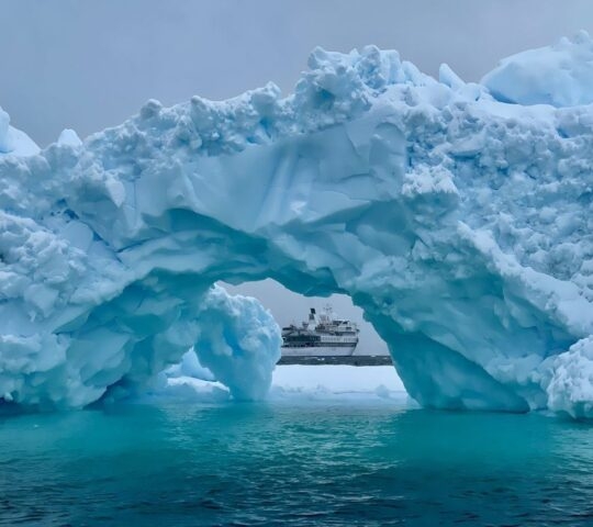 An expedition cruise ship in Antarctica as seen through the arch of an iceberg.