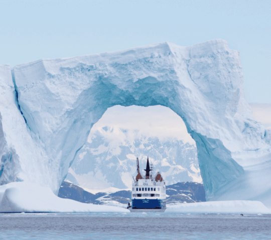 Expedition cruising ship sailing between the arch of a giant iceberg.