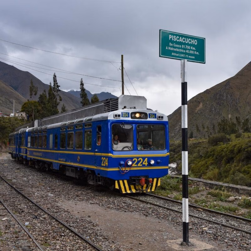 A blue and yellow train on tracks in a mountain valley, passing a sign for "PISCACUCHO," often a part of luxury Machu Picchu tours.