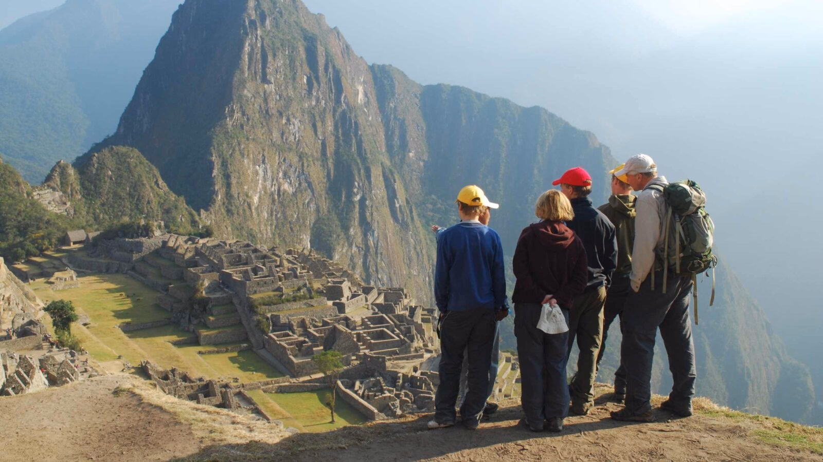 : Five tourists overlooking the stone ruins of Machu Picchu with Huayna Picchu mountain in the background.