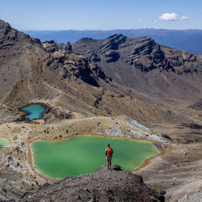 A woman stands looking over a bright green crater lake and volcanic landscapes in Tongariro, New Zealand