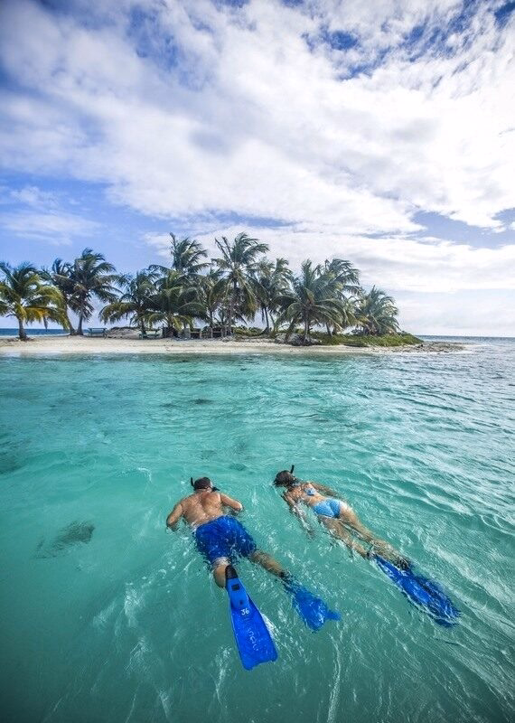 A man and woman snorkel in bright turquoise water toward a small, palm-covered island under a cloudy blue sky. Highlight of luxury Belize tours.