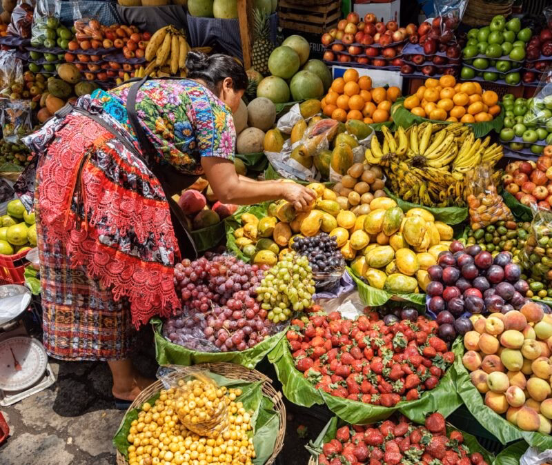 Woman wearing colorful traditional attire arranging mangoes at a vibrant outdoor market with strawberries, grapes, and bananas.