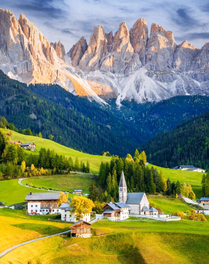 A picturesque village with chalets and a church nestled in a green valley below towering grey peaks.