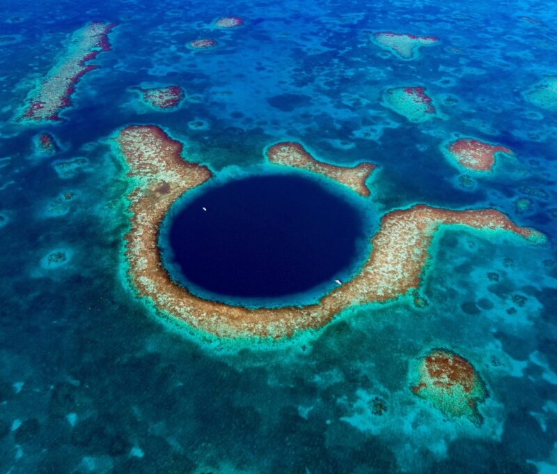 Aerial view of a large circular deep blue sinkhole in the middle of a turquoise coral reef in the ocean.