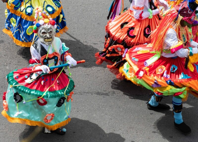 People in bright, ruffled carnival costumes and a monster mask dancing on a grey asphalt street during a parade.