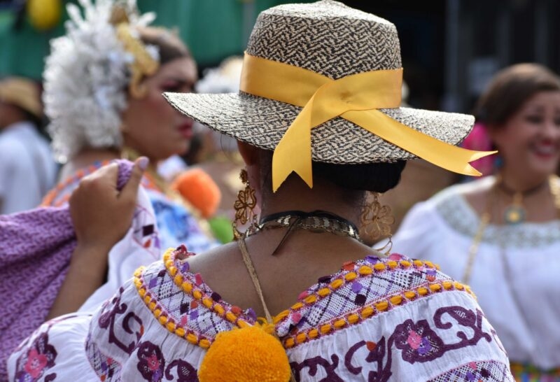 Close-up from behind of a woman in a white embroidered dress and straw hat with a yellow ribbon at a parade.