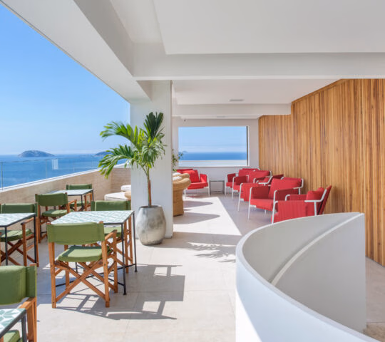 An open-air terrace with white ceiling and floor. On the right are red lounge chairs, on the left are green chairs and dining tables. In the background is a clear glass railing and beyond is the blue sea and sky with a few islands in the distance.