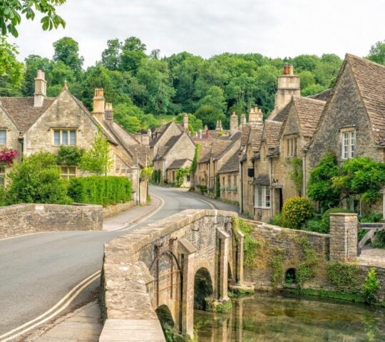 A stone bridge over a small stream leading into a village of historic stone cottages nestled among green trees.