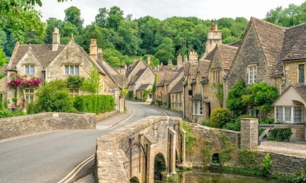 A stone bridge over a small stream leading into a village of historic stone cottages nestled among green trees.