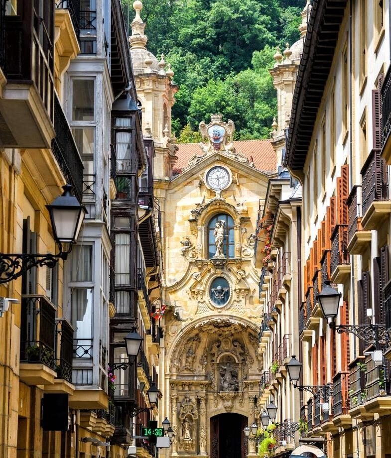 A narrow street leading to the ornate Basilica of Saint Mary in San Sebastián and rolling green hills dotted with stone villages across the Cotswolds.