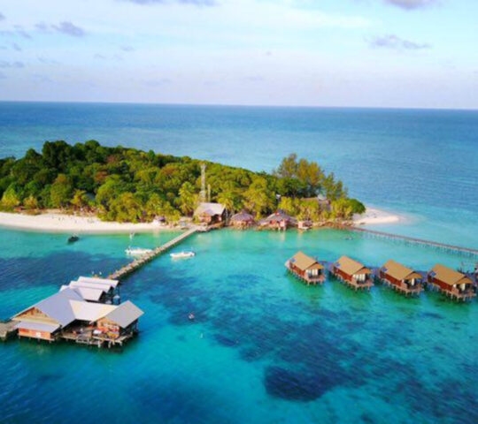 Aerial shot of a resort on a tiny island surrounded by white sand and turquoise water and covered in dark palms. A long pier extends from one side with a building over the water, and on the other side, a row of smaller villas run perpendicular to the beach over the water.