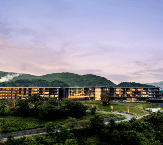 Wide view of a modern hotel on a hilltop at sunset with green hills behind it