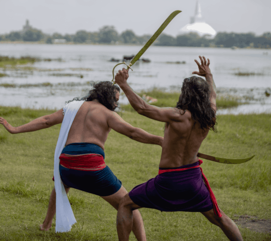 Two martial artists practising the ancient martial art of Angampora beside an expanse of water in Sri Lanka.