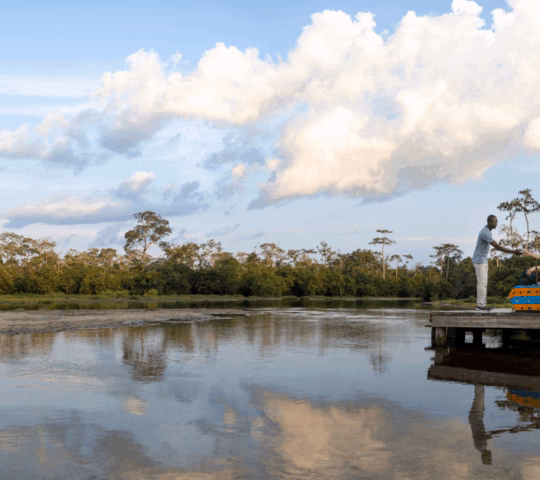 A man serving a woman who is seated on a wooden platform raised above the waters of the Lango baï