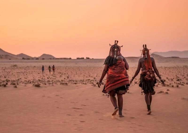 A group of children play in the foreground while a Himba woman with a young child wrapped onto her back walks beside another Himba woman.