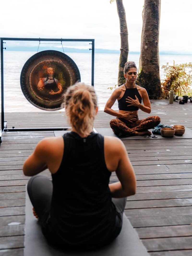Two women in a meditative seated pose on a wooden platform with the ocean behind them