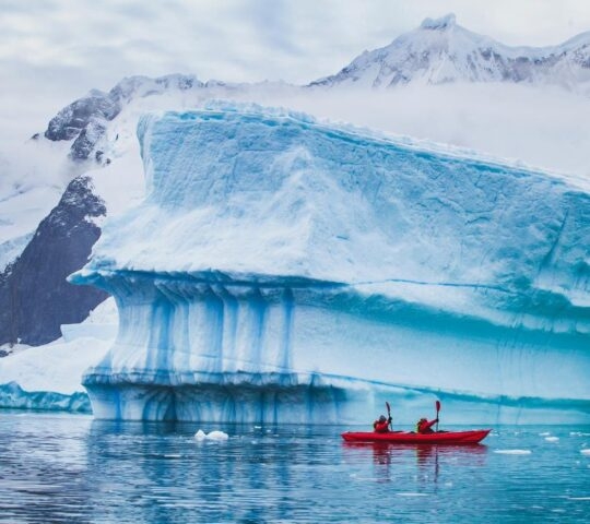 A couple in a red kayak paddling past an iceberg in Arctic waters