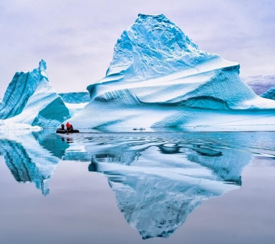 A zodiac boat approaching an iceberg reflected in still water