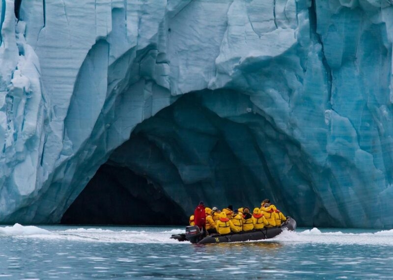A group of people on a Zodiac excursion in the Arctic, cruising past a cave carved into a giant glacier.