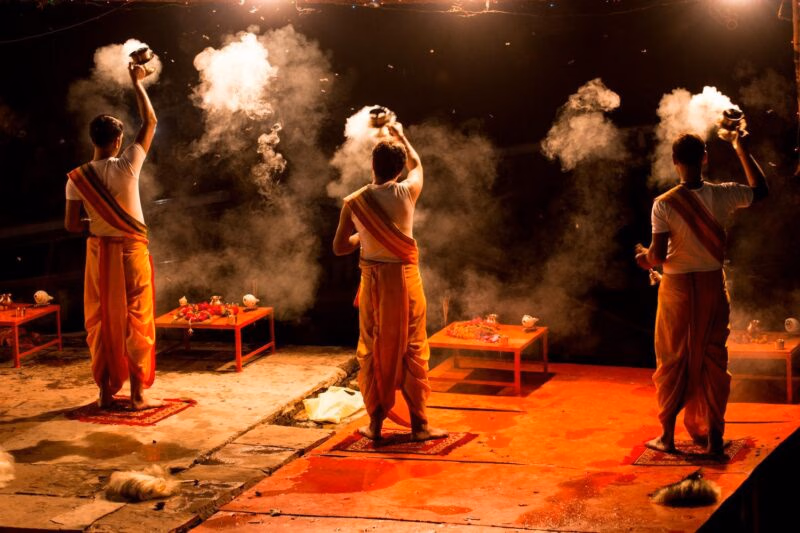 Three monks dressed in orange holding flaming lanterns in the darkness as part of an Aarti ceremony in Varanasi