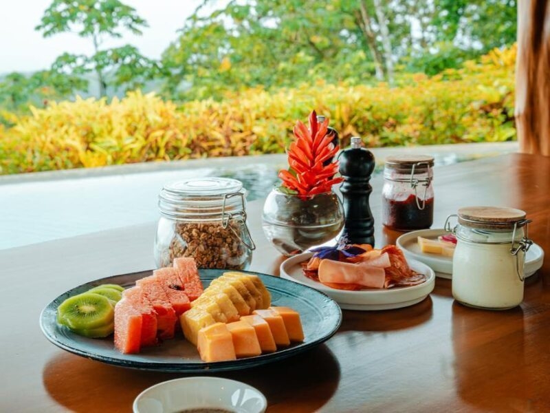 A wooden table with a fruit platter and breakfast plates in front of a pool and trees.