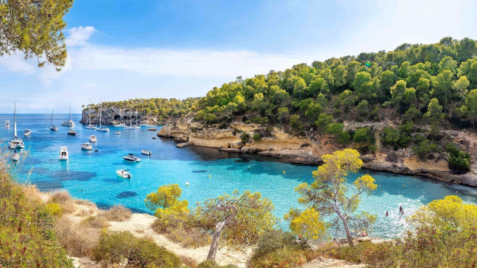 View of a bay with boats floating in turquoise water, surrounded by rocks and trees in Mallorca, Spain