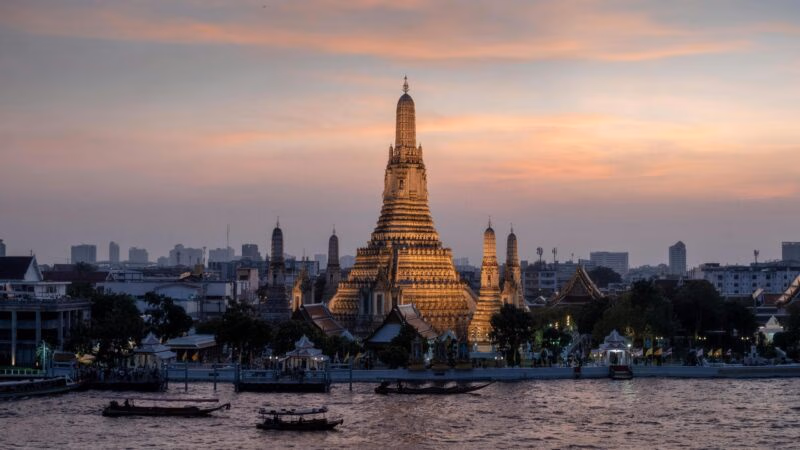 Illuminated Wat Arun temple at sunset overlooking the Chao Phraya River in Bangkok.