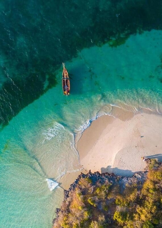 Top-down aerial view of a wooden boat in clear turquoise water next to a white sand tropical beach.