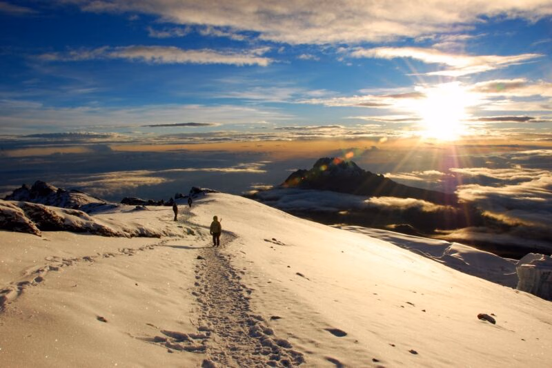 Hikers walk on a snow-covered path on Mount Kilimanjaro at sunrise with clouds below the peak.