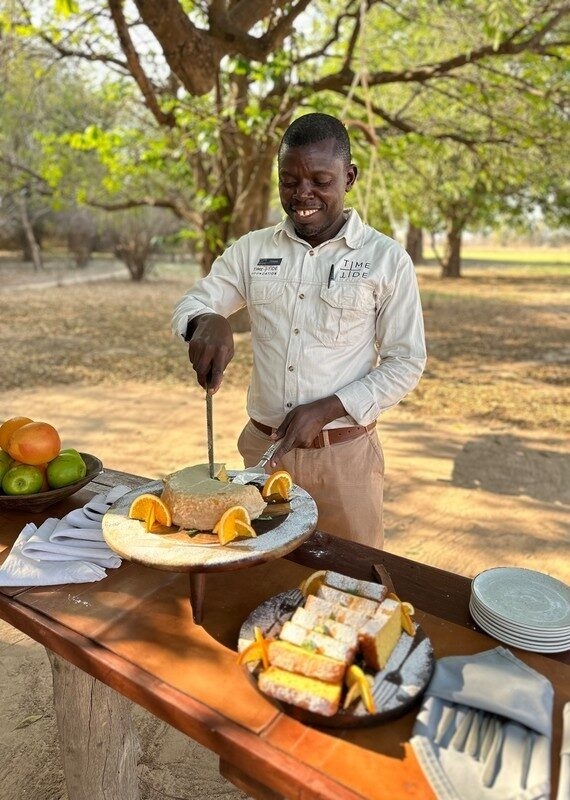 A man in a tan uniform cutting a cake on a wooden table set with fruit and pastries outdoors.