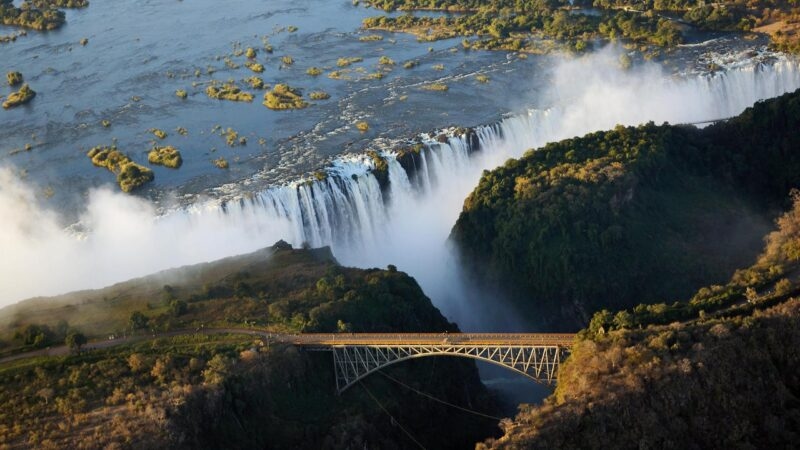 High-angle view of a steel arch bridge over a river gorge next to large waterfalls and mist.
