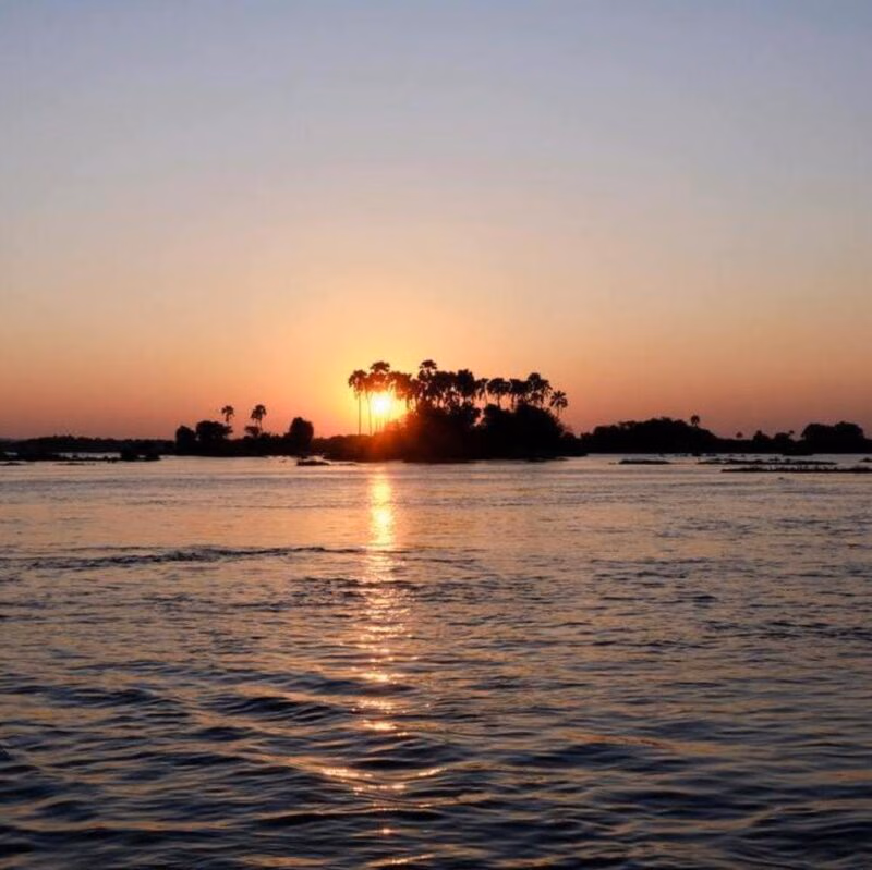 A glowing sun setting behind a small island with palm trees in the middle of a wide river.