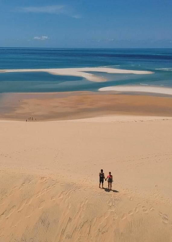 Two people standing on a high sand dune overlooking a tropical coastline with turquoise water and sandbars.