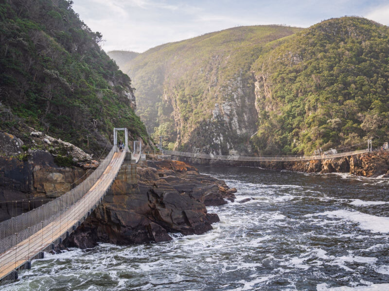 A narrow suspension bridge stretches across a rocky gorge over churning water between steep, forested hills.