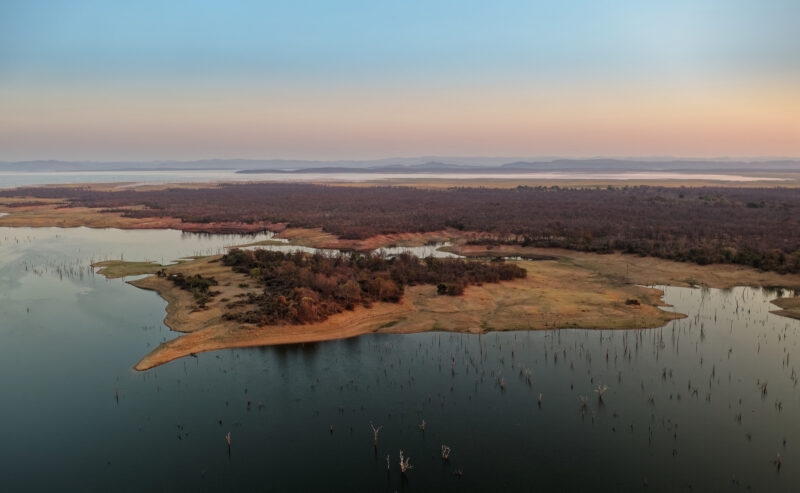 High-angle wide shot of Lake Kariba's shoreline and calm water under a soft pink and blue sunset sky.