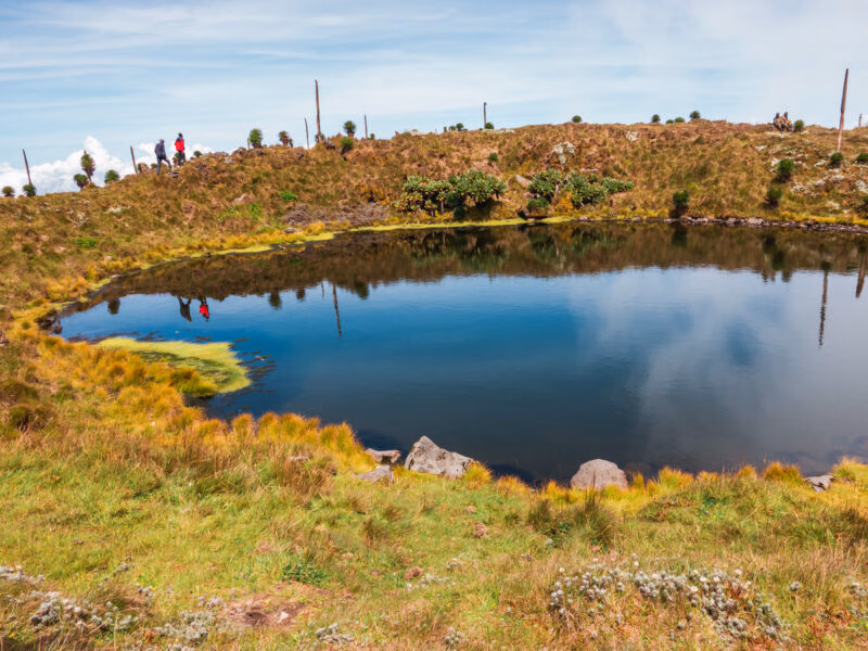 A still crater lake reflecting the blue sky, surrounded by grassy banks and hikers on the ridge.