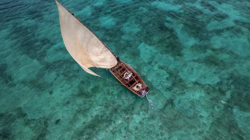 Top-down view of a traditional dhow boat sailing over a clear coral reef in the Bazaruto Archipelago.