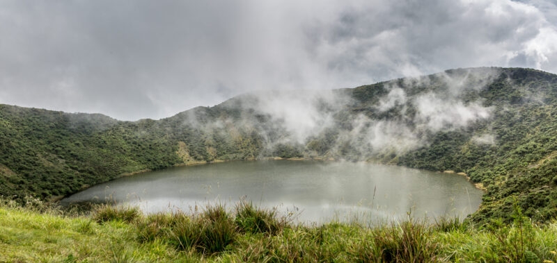 A wide crater lake tucked into green mountains with thick white mist rolling over the water in Virunga National Park.