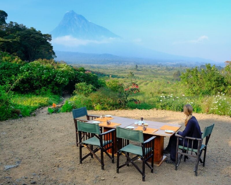 An outdoor dining table with orange placemats facing a large blue volcano shrouded in low clouds in Virunga National Park.
