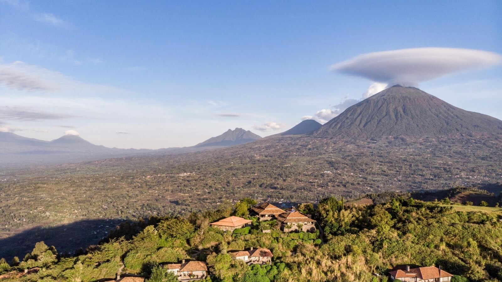 Aerial view of a luxury lodge on a lush green hilltop with large volcanoes in the background of Virunga National Park.