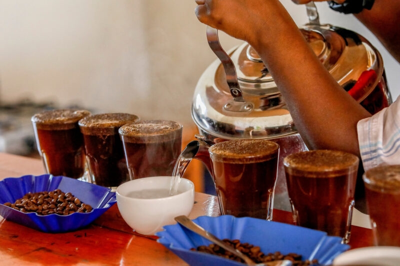 Close-up of coffee being prepared with a silver kettle and glasses of brew at a Kigali coffee tasting.