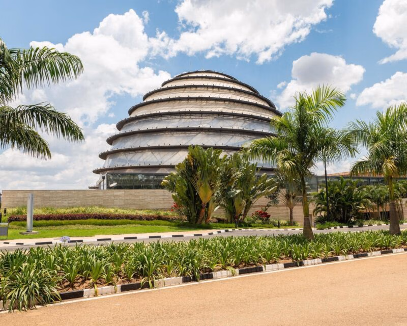 The modern, tiered dome of the Kigali Convention Centre behind tropical greenery and a bright sky.