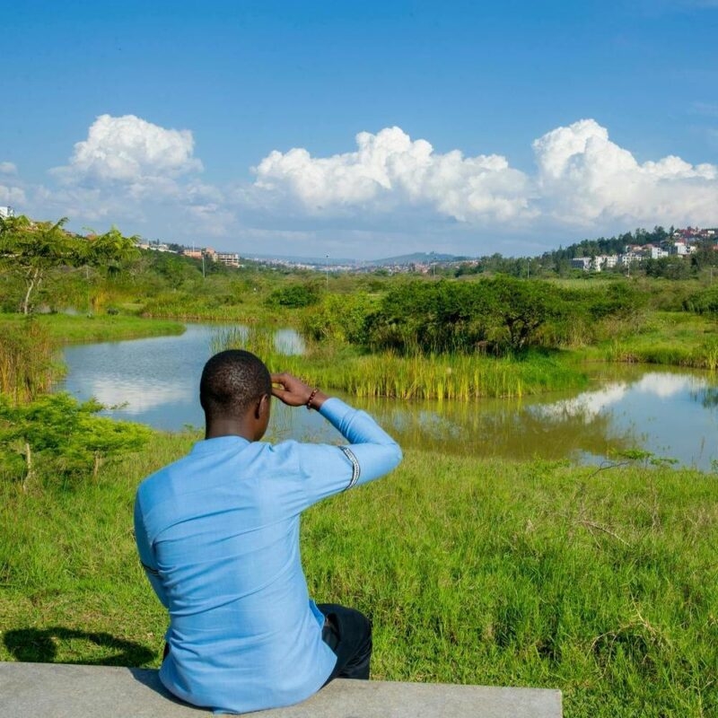 A man in a blue shirt sits on a bench looking out over a calm lake and wetlands in Kigali.