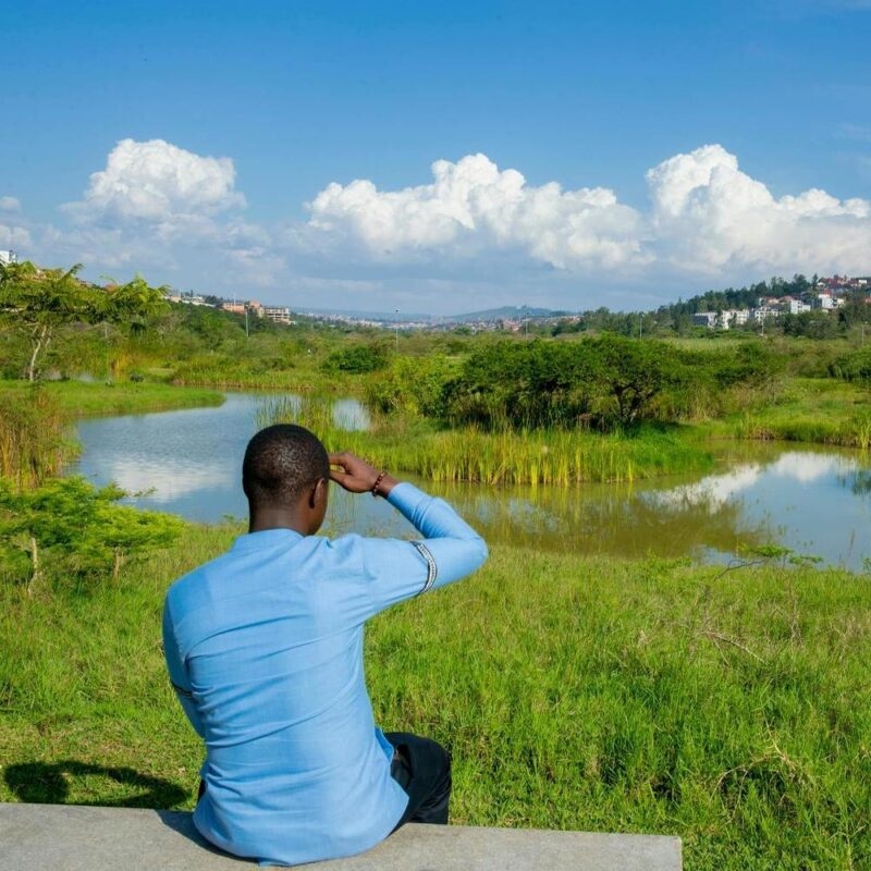 A man in a blue shirt sits on a bench looking out over a calm lake and wetlands in Kigali.