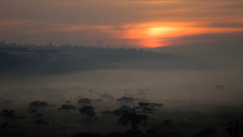 A glowing orange sun dips below the horizon over a misty, tree-filled valley in Kigali at dusk.