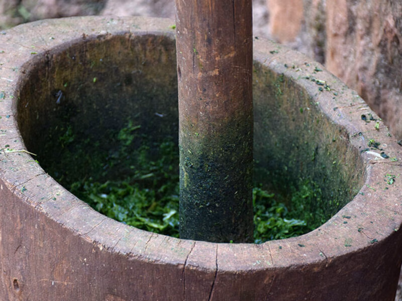 A wooden pestle mashing green leafy vegetables inside a traditional deep wooden mortar in Kigali.