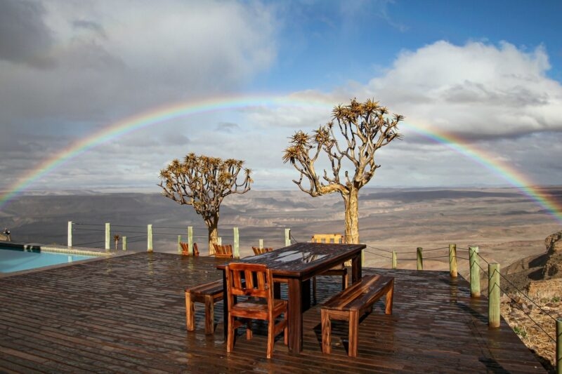 Outdoor wooden table and benches on a deck with Quiver trees and a vibrant rainbow over the canyon.