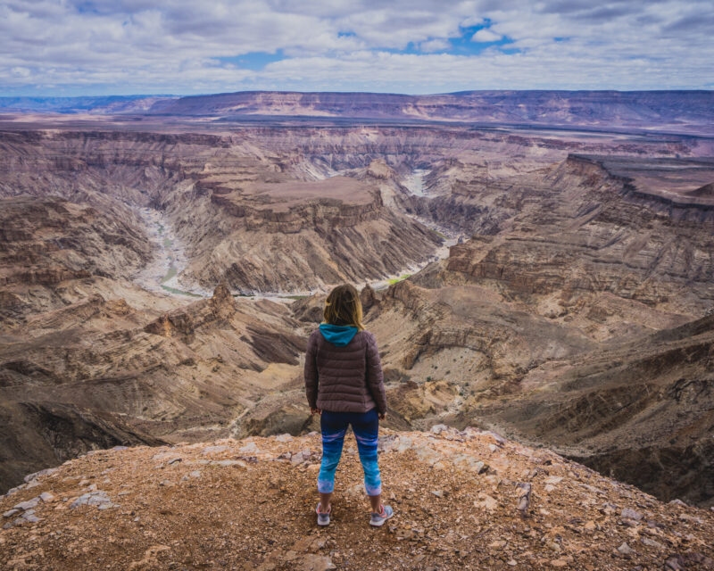 Hiker overlooking the deep, rocky ravines and winding paths of Fish River Canyon under a cloudy sky.