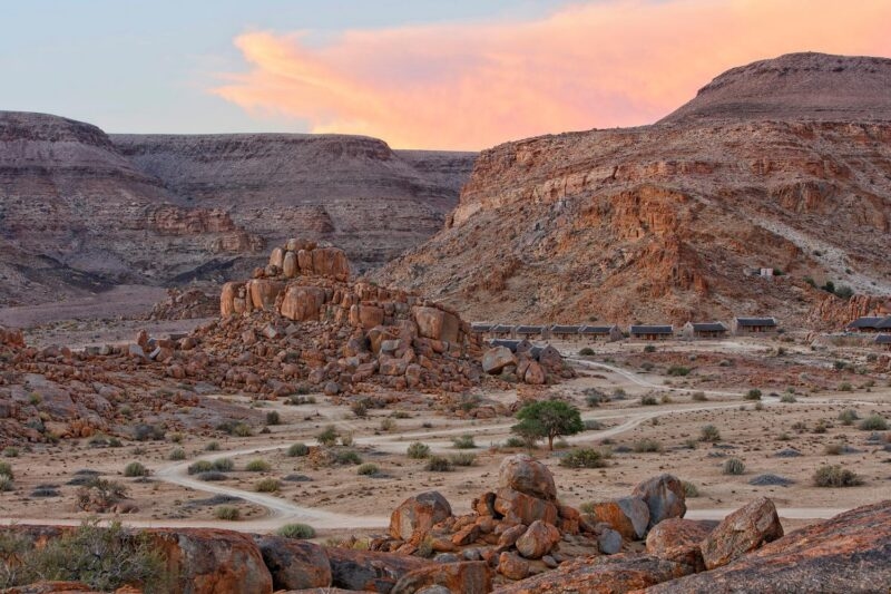 Desert lodge structures and a dirt path among massive orange rocks under a soft pink sunset sky.